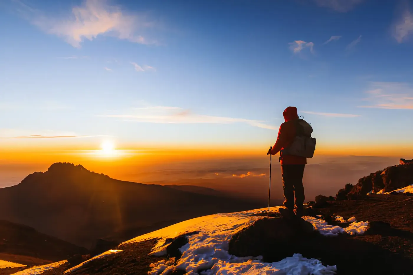 A hiker wearing a backpack stands on a snowy mountain ridge at sunrise, overlooking a vast landscape glowing with orange and golden light beneath a clear blue sky.